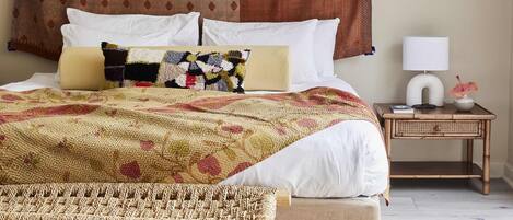 Uma cama bem arrumada com um cobertor estampado, uma mesinha de cabeceira com uma luminária e uma pequena planta, e um ar-condicionado fixado na parede.