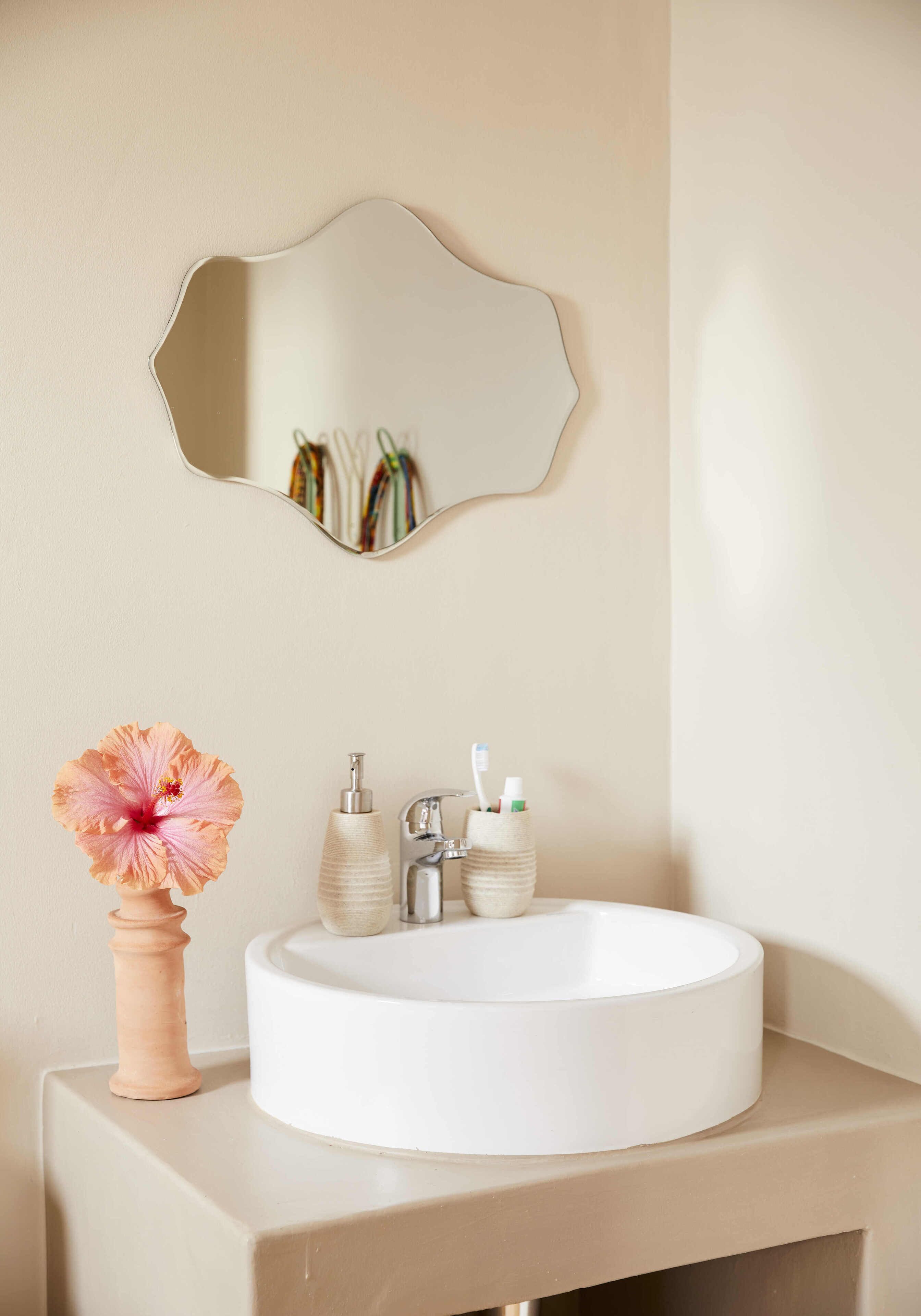 A bathroom with a round white sink, a mirror, and a vase with a flower.