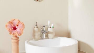 A bathroom with a round white sink, a mirror, and a vase with a flower.