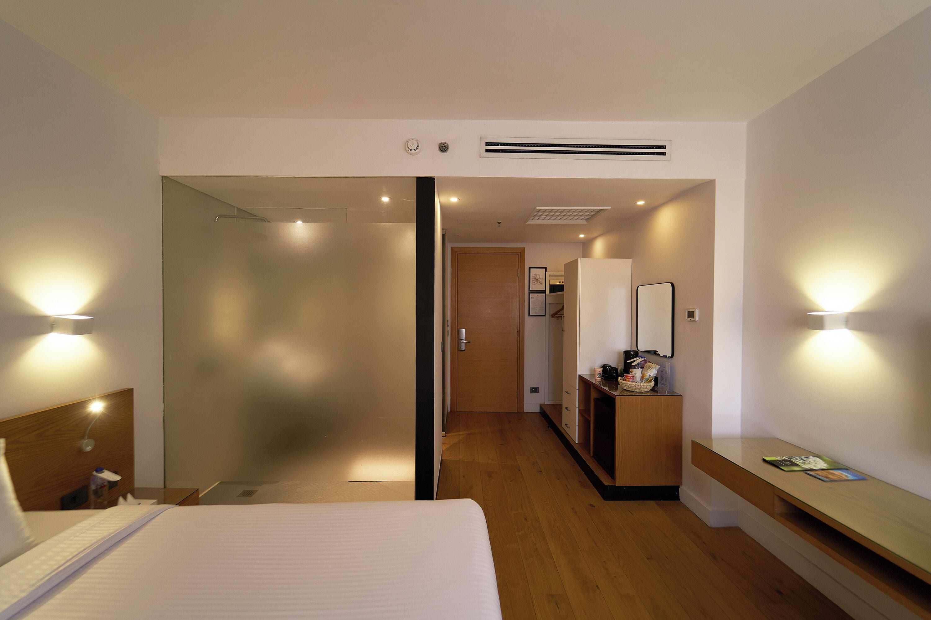 Una habitación de hotel con cama, mesita de noche, lámpara y cómoda de madera con espejo.