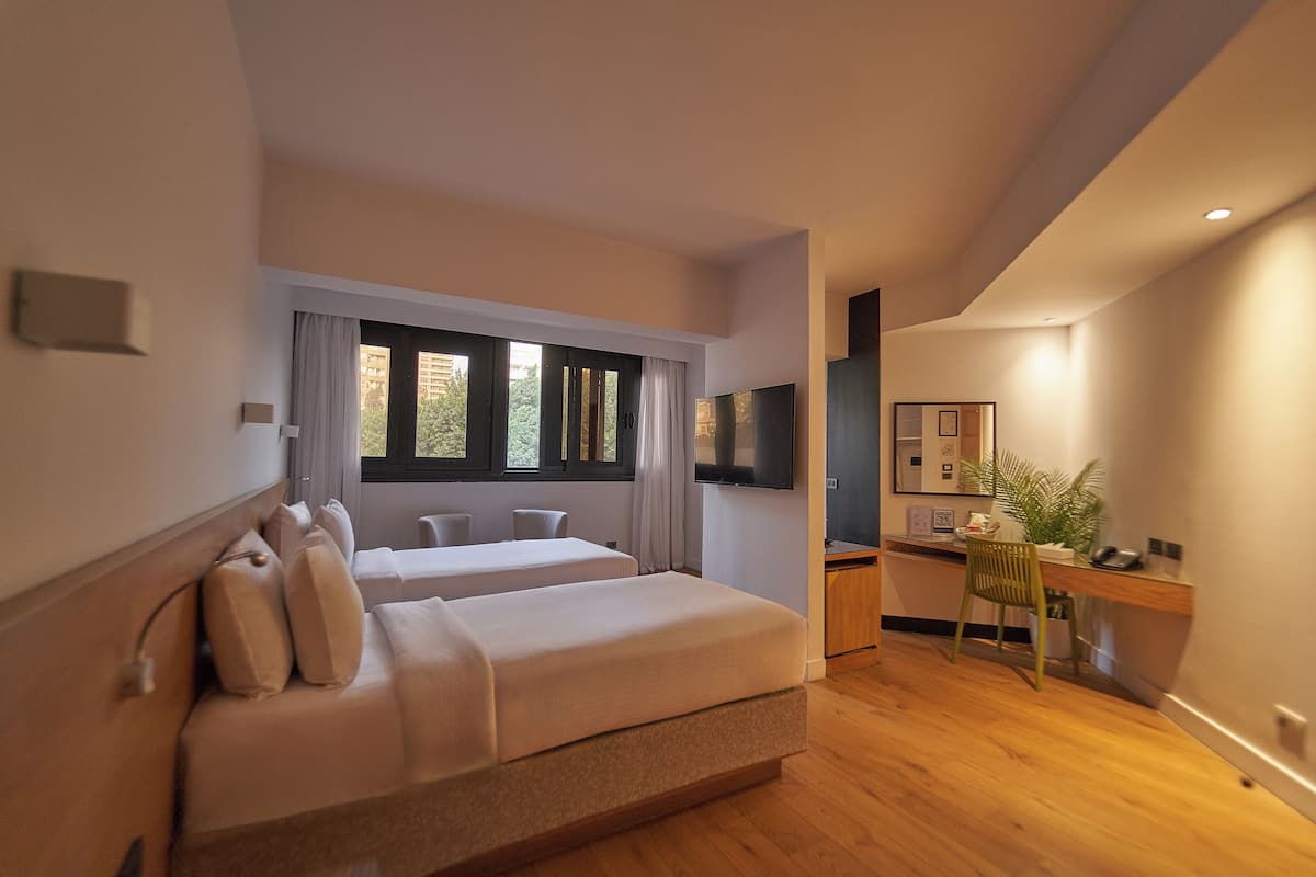 A modern hotel room with two beds, a desk, a chair, a TV, and a large window.
