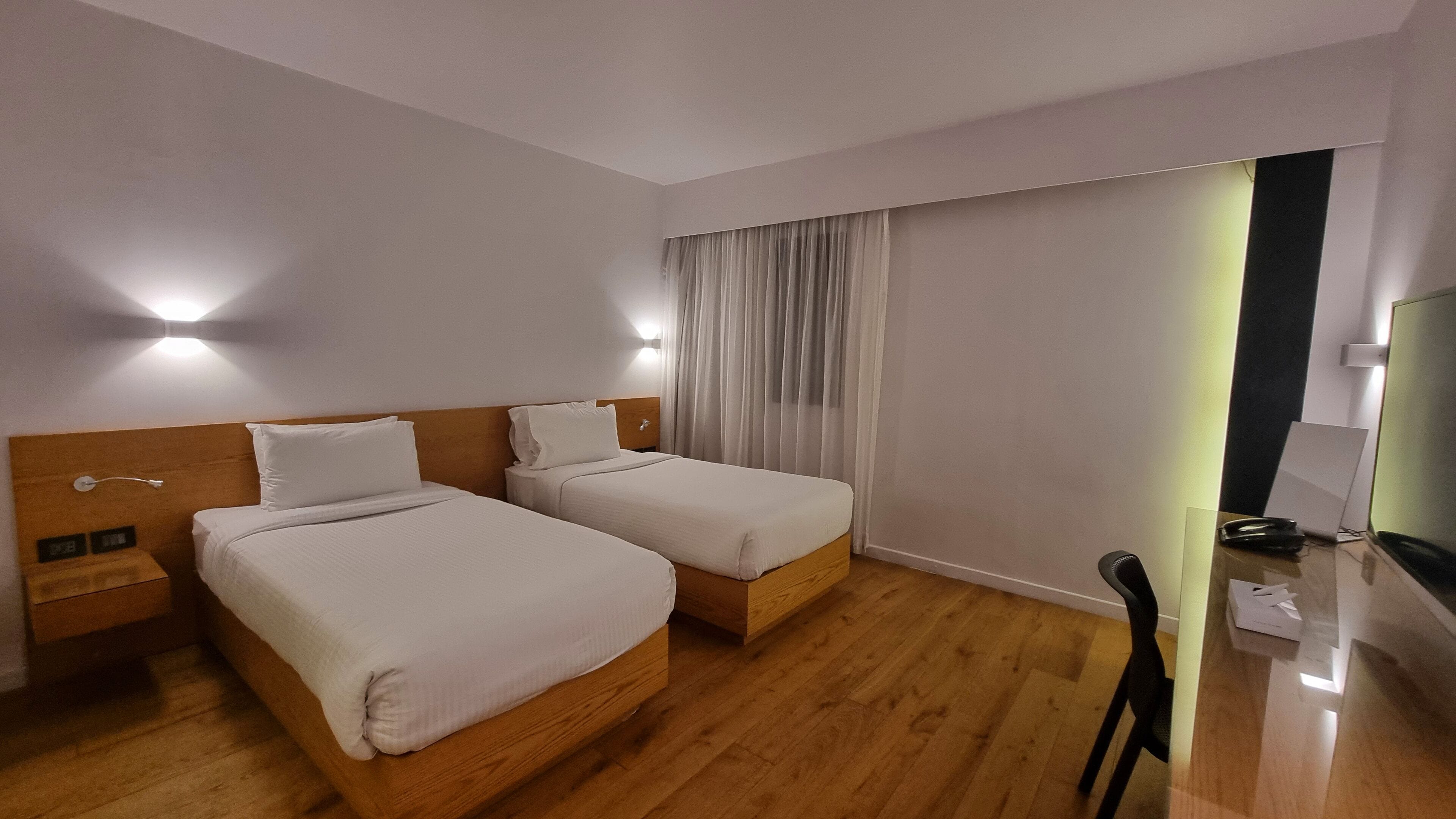 Hotellihuone, jossa on kaksi sänkyä, puulattia, työpöytä tuolilla, televisio ja verho.