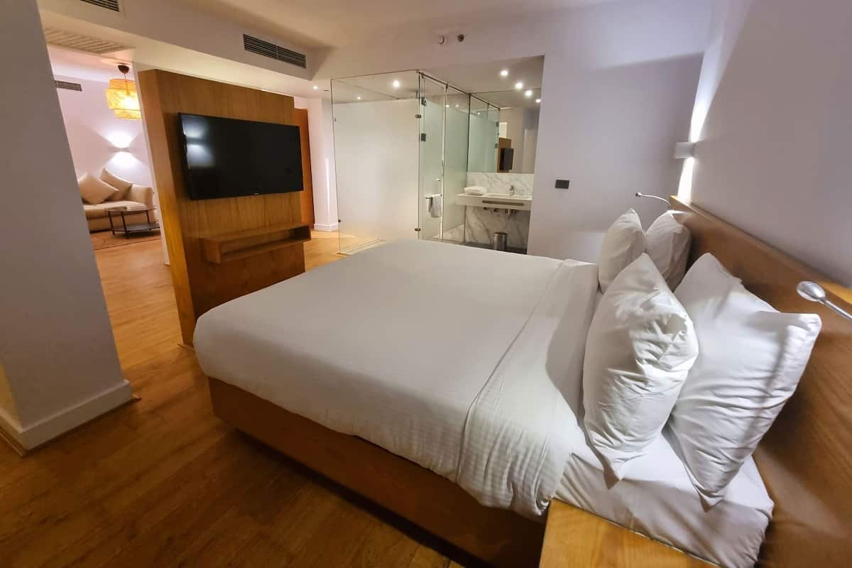 A modern hotel room with a large bed, a television, a bathroom with a mirror, and a seating area with a sofa.