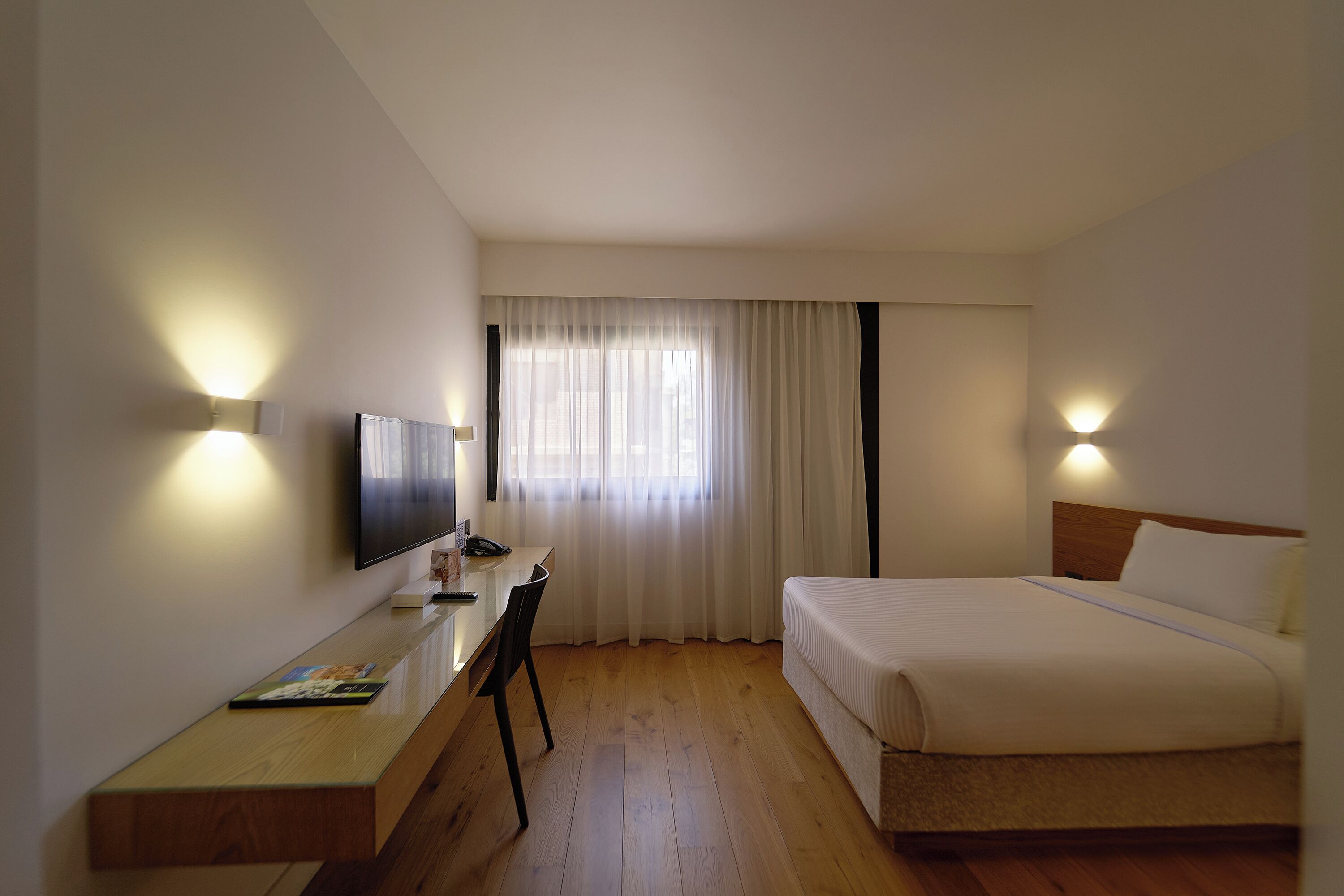 Une chambre d’hôtel comprenant un lit, un bureau avec une chaise, un téléviseur et deux lampes fixées au mur.