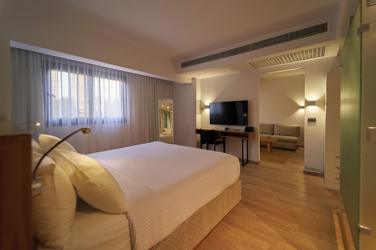 A hotel room with a large bed, a desk with a flat-screen TV, a sofa, and a dining table.