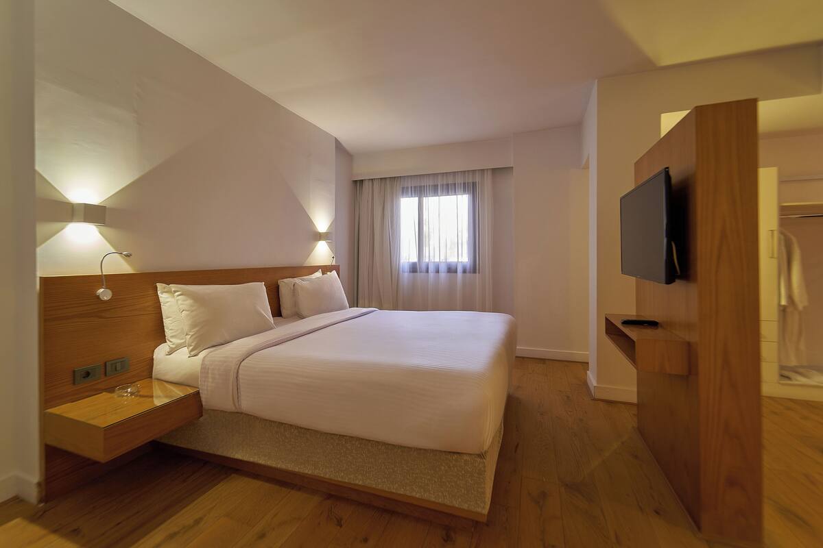A hotel room with a wooden bed, bedside tables, a flat-screen TV, and a large window with curtains.
