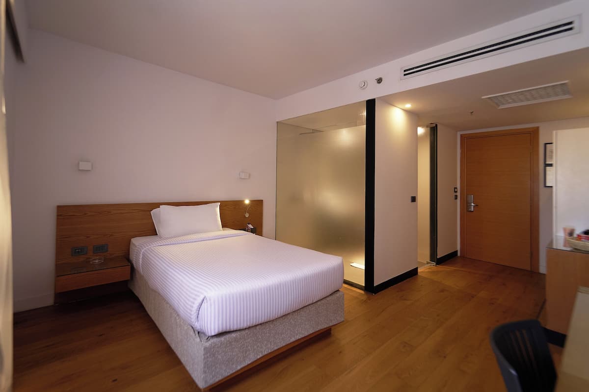 A modern hotel room with a wooden bed, a nightstand, a desk, and a door.