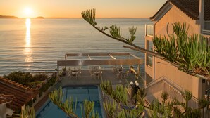 A poolside area with a view of the sea at sunset, outdoor seating, and a building with a tiled roof. - Denise Beach Hotel (Zakynthos)