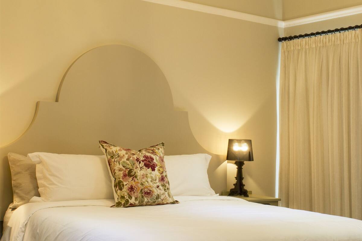 Una cama bien hecha con sábanas blancas, un cojín floral y una lámpara de noche.