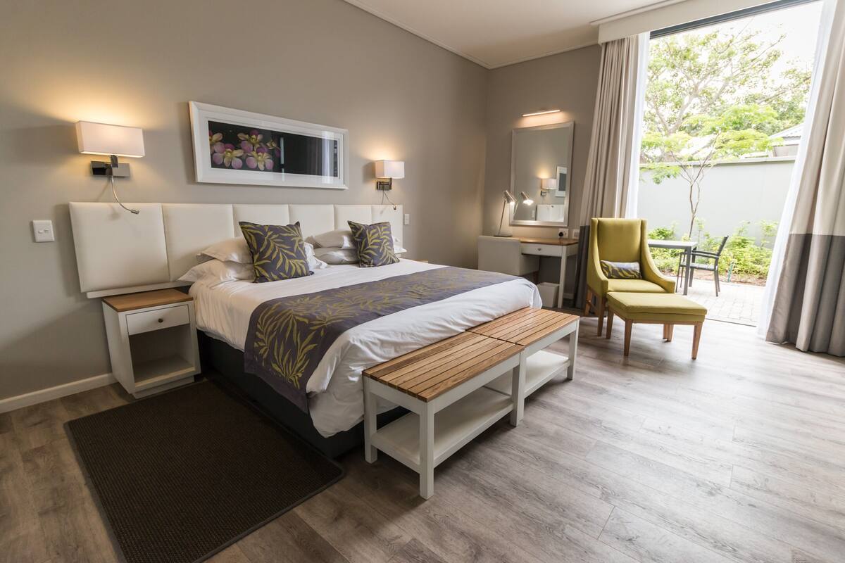 Habitación de hotel con cama, mesitas de noche, un banco, una silla, un escritorio y un ventanal grande.