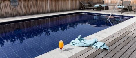 Una piscina con cubierta de madera, sillas de descanso y un vaso de jugo de naranja.