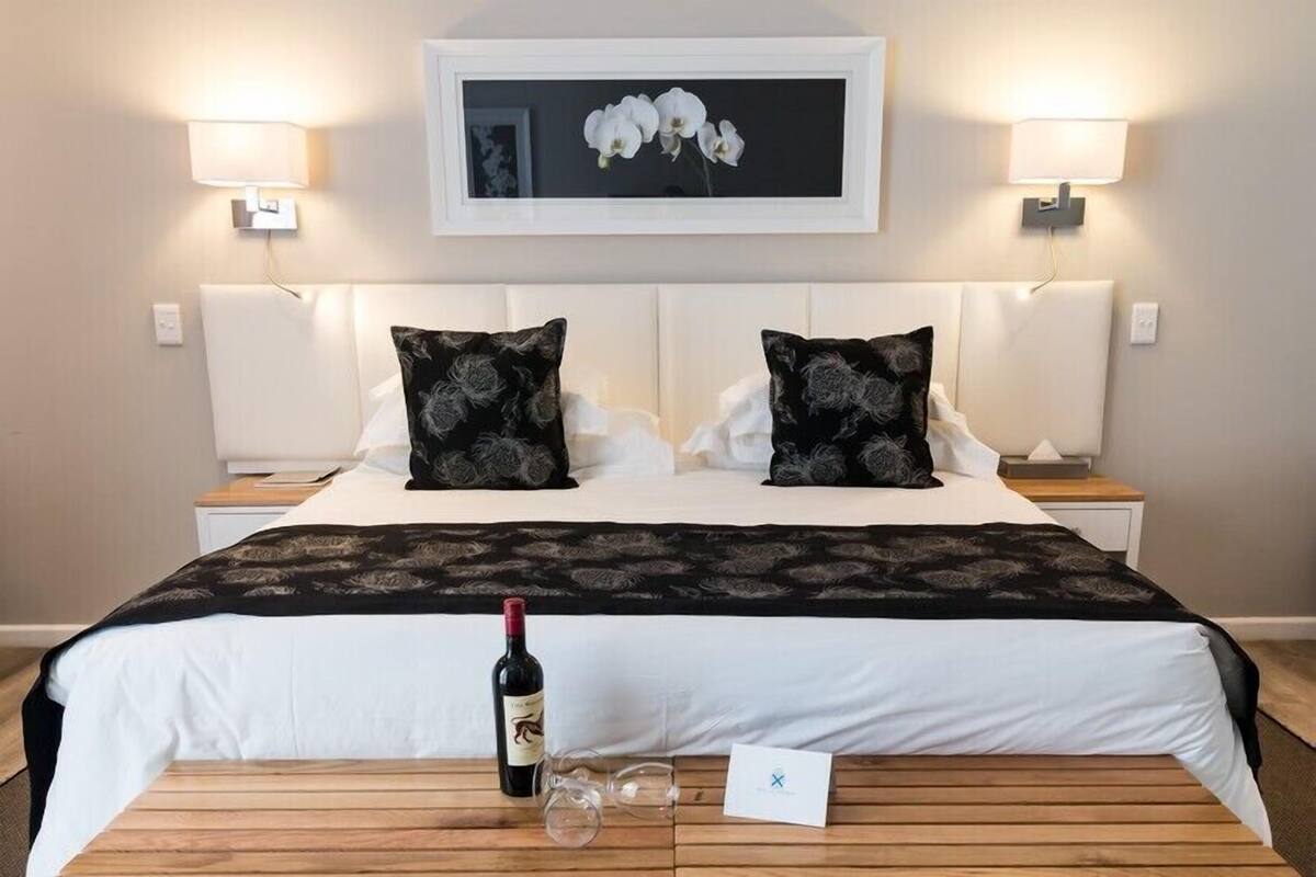 Habitación de hotel con una cama grande, dos mesitas de noche, un cuadro enmarcado en la pared y una botella de vino sobre la cama.