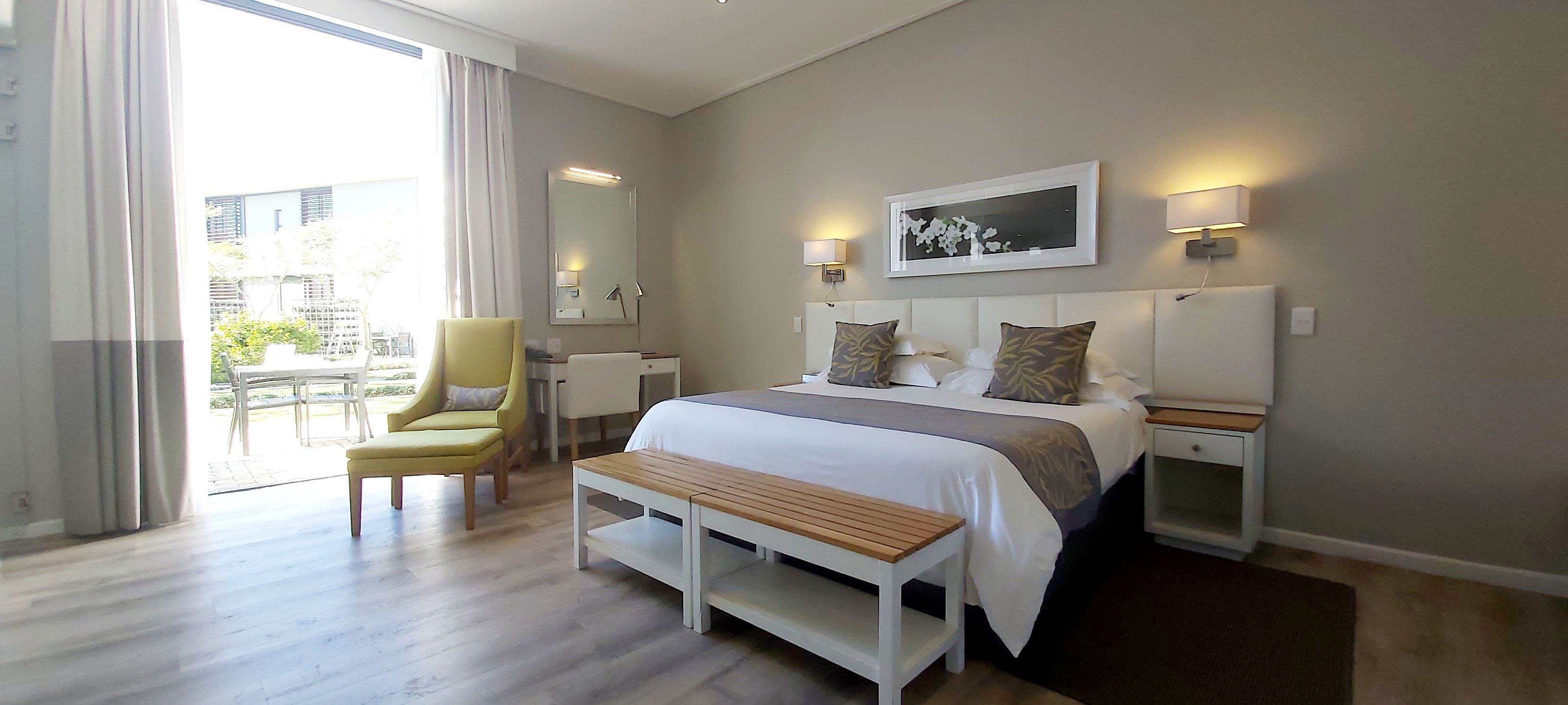 Une chambre d’hôtel avec un lit, des tables de chevet, une chaise, un banc, une grande fenêtre et une vue sur l’extérieur.