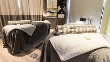 Two beds with dark bedding and plaid-patterned pillows, a bedside table, and a wall-mounted lamp are featured in this stylish room.