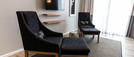 Un salon moderne avec deux fauteuils noirs, une table basse en bois, un téléviseur à écran plat et une œuvre d’art encadrée accrochée au mur.