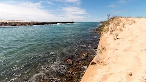 Paisaje costero con aguas azules cristalinas, una playa de arena y un rompeolas rocoso.