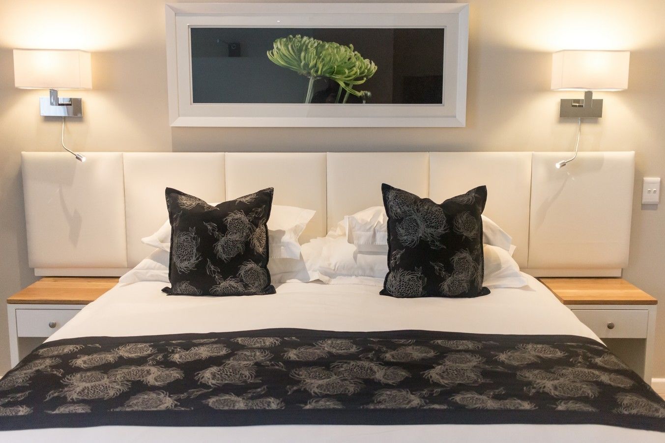 Una cama cuidadosamente tendida con una colcha blanco y negro estampada, dos almohadas negras con un diseño plateado, y un cuadro con una planta colgado en la pared de arriba.
