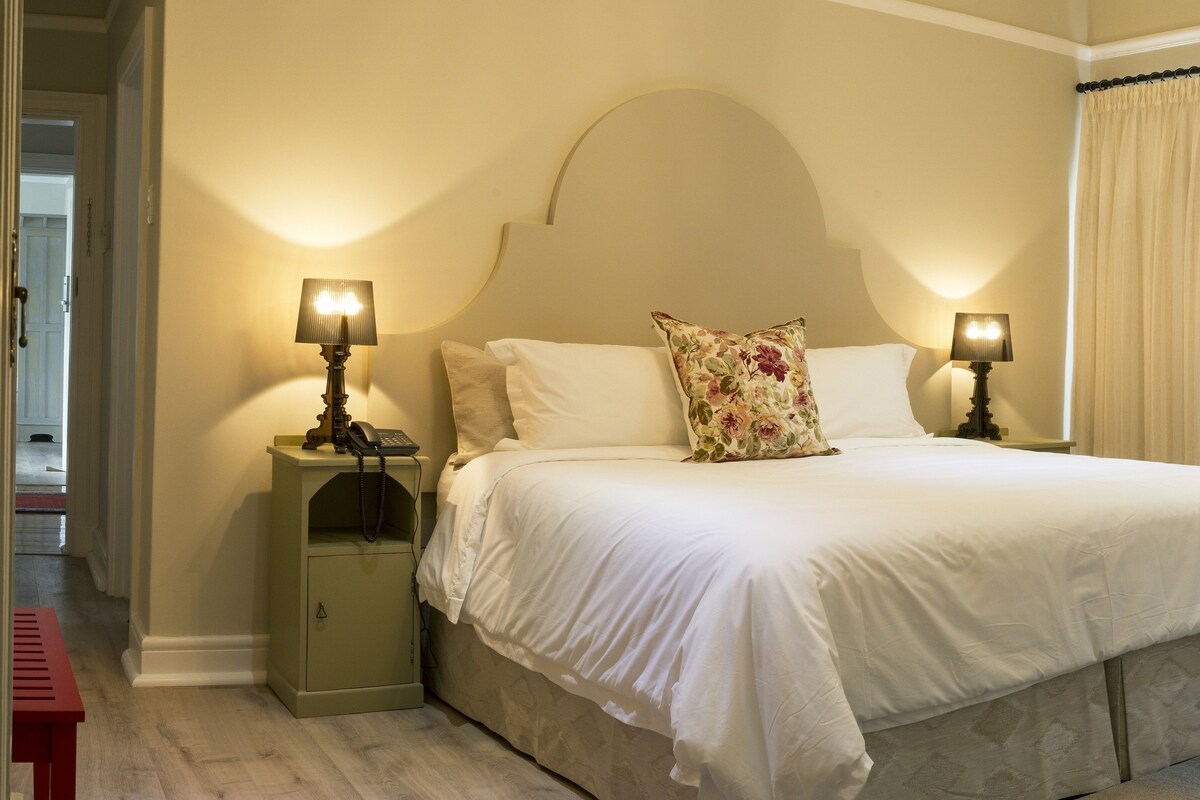 A neatly made bed with white linens, two bedside lamps, a floral pillow, and a small bedside table with a phone.