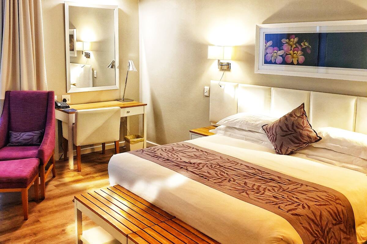 Una habitación de hotel con cama, mesita de noche, silla, escritorio, lámpara y un espejo grande.