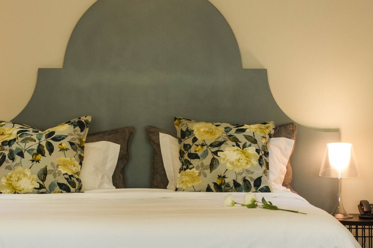 Una cama bien hecha con almohadones florales, un cabecero grande, lámparas de noche y cuadros en la pared.