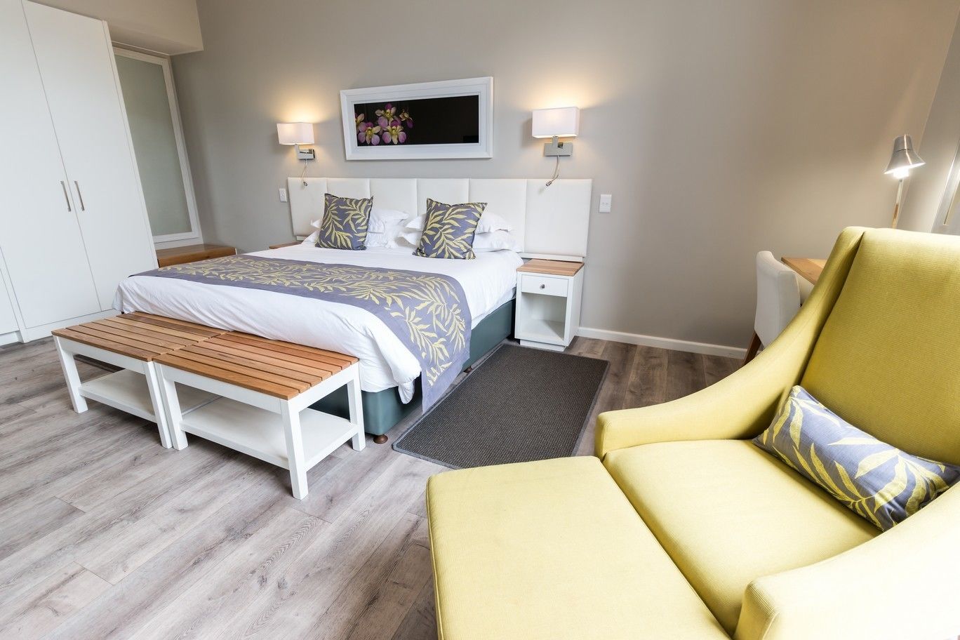 A bedroom with a bed, bedside tables, a bench, a chair, and a wall-mounted TV.