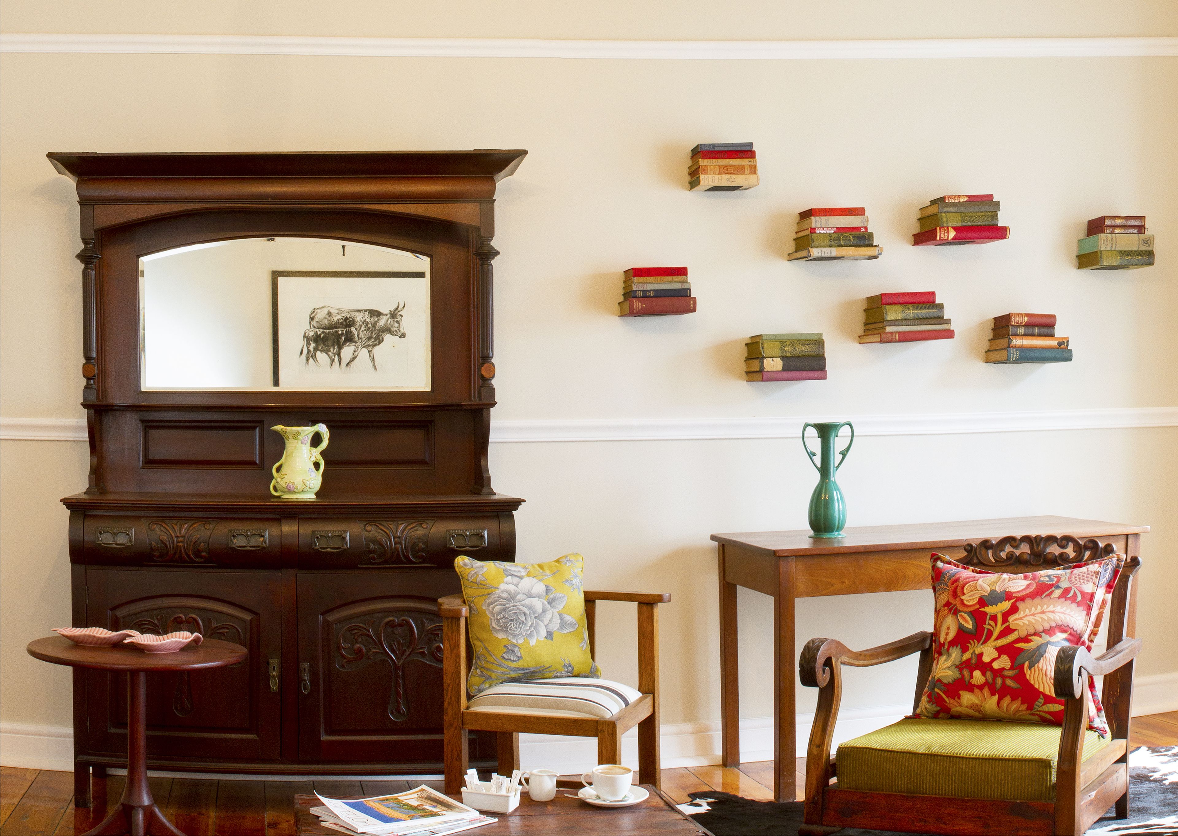 A room with a wooden cabinet, a chair with a floral cushion, a small table with a cup, and a wall with stacked books.