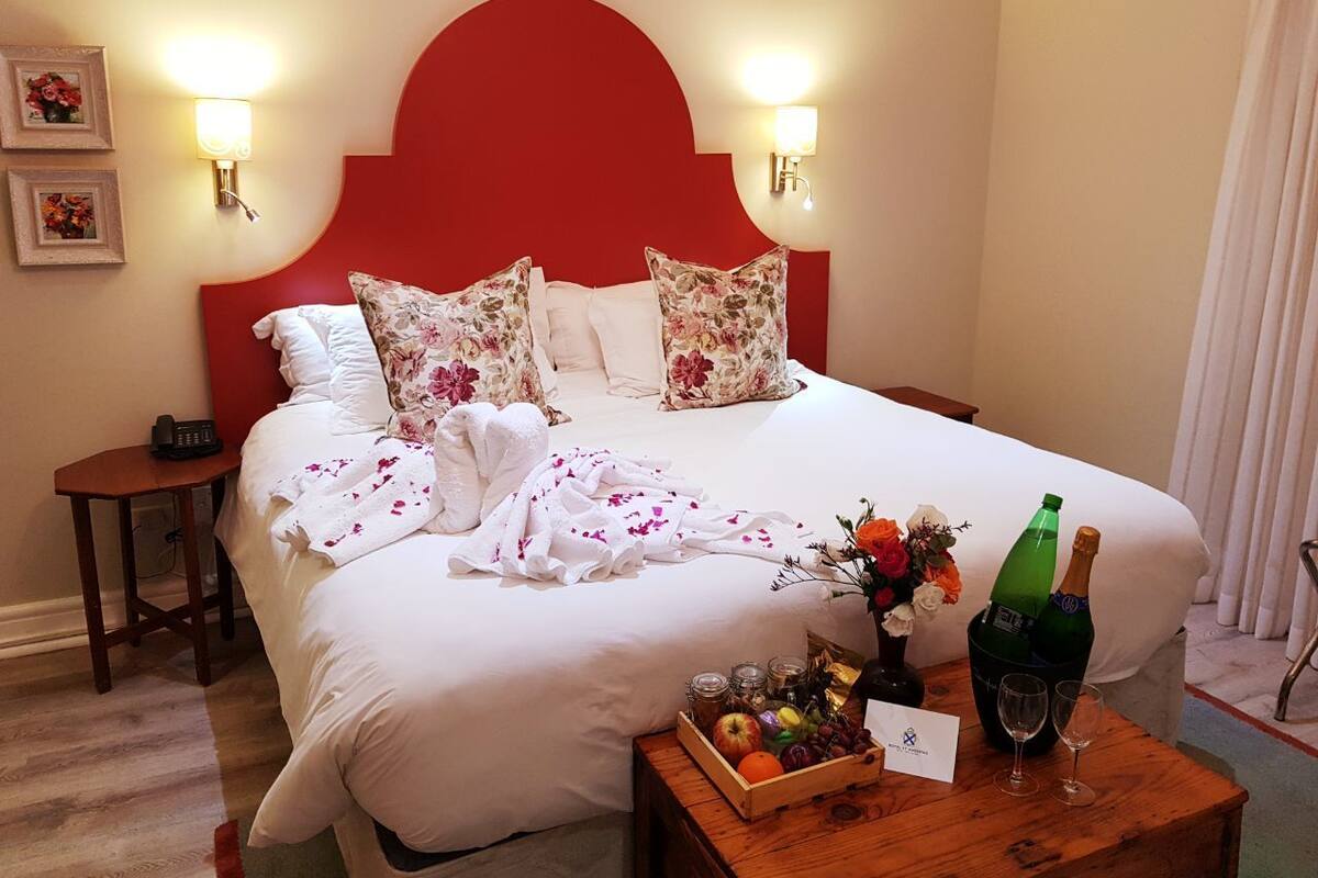 Una habitación de hotel con una cama grande, almohadas estampadas con motivos florales, una mesita de noche con una botella de champán y una bandeja de fruta.