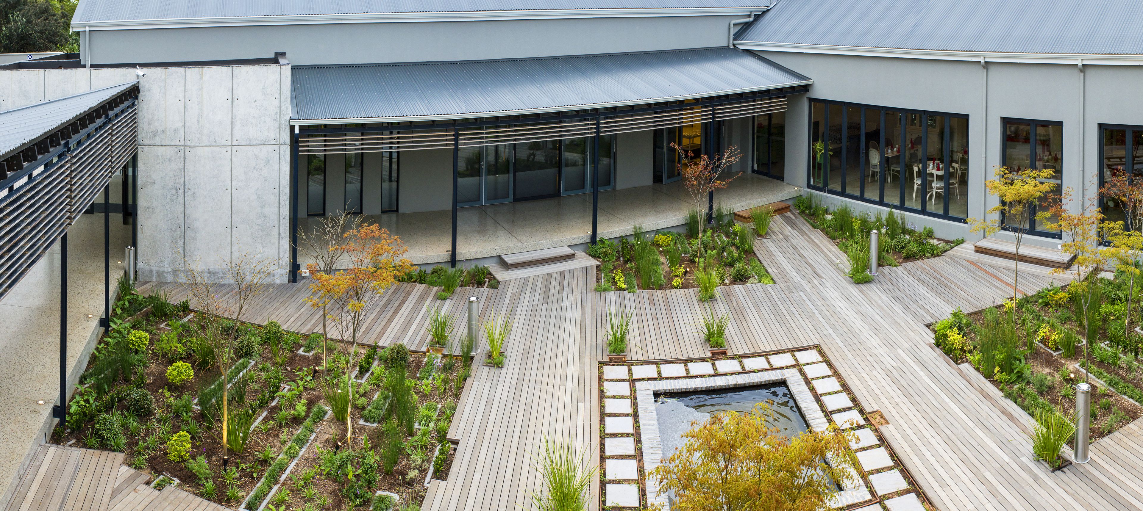 A modern building with a covered outdoor area, a garden with various plants, and a small water feature.