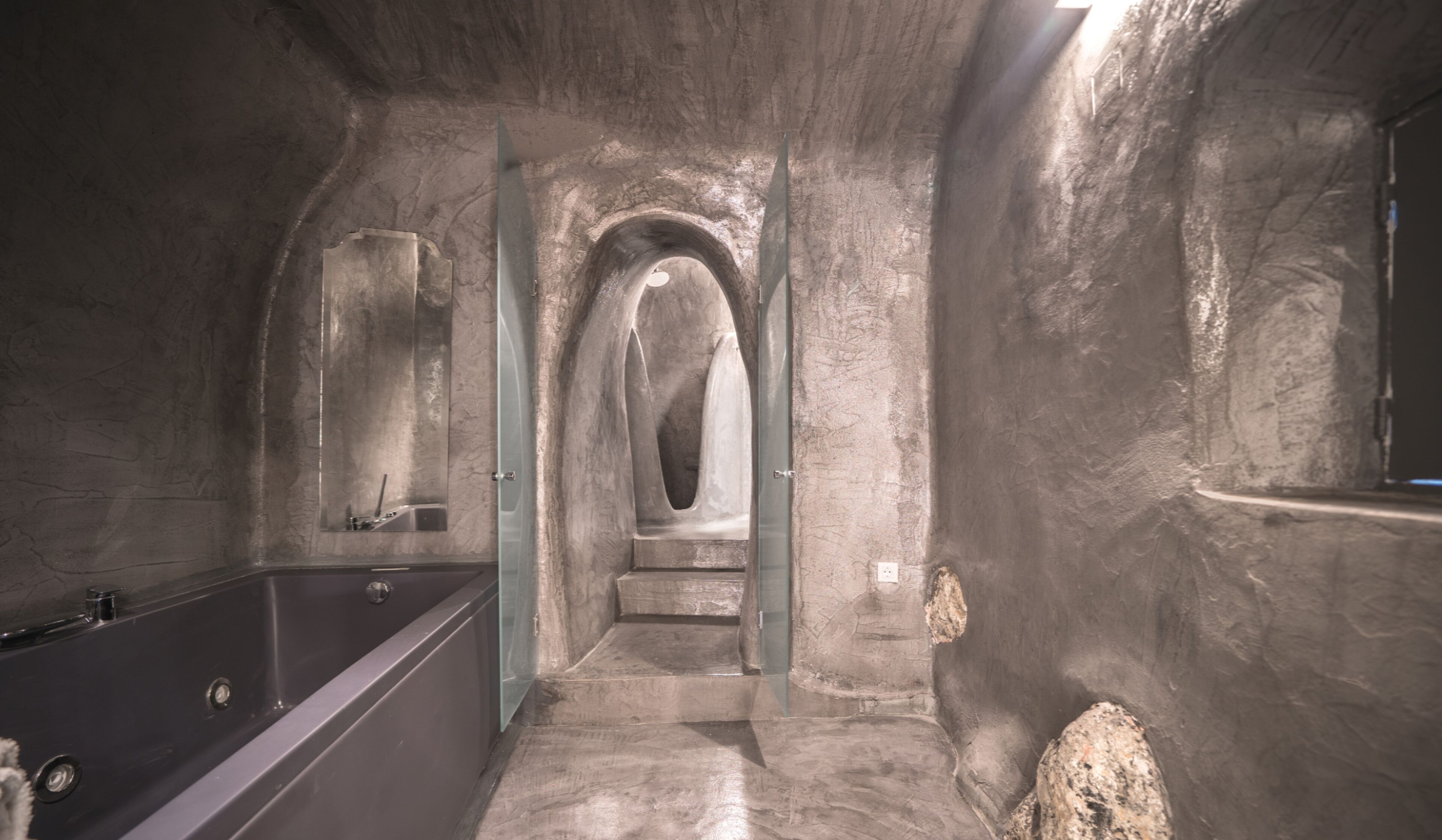 A bathroom with a bathtub, a sink, and a mirror. There is a stone wall and a stone archway.