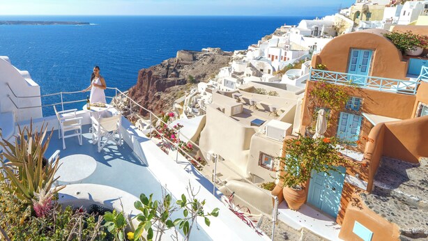Vista costera con edificios blancos, una persona con vestido blanco y mar azul cristalino.