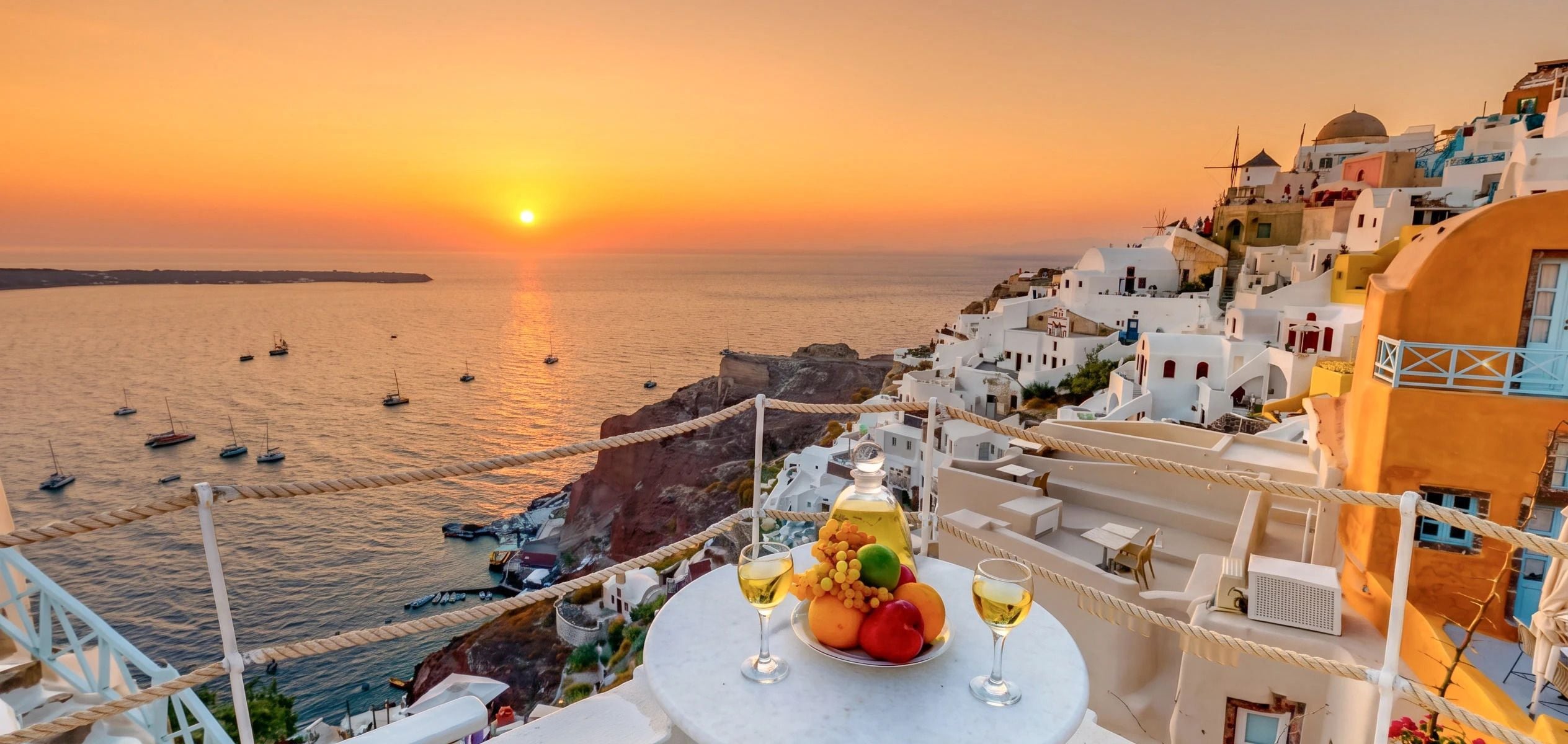 A seaside view with a sunset, boats on the water, and a table with fruit and wine glasses.