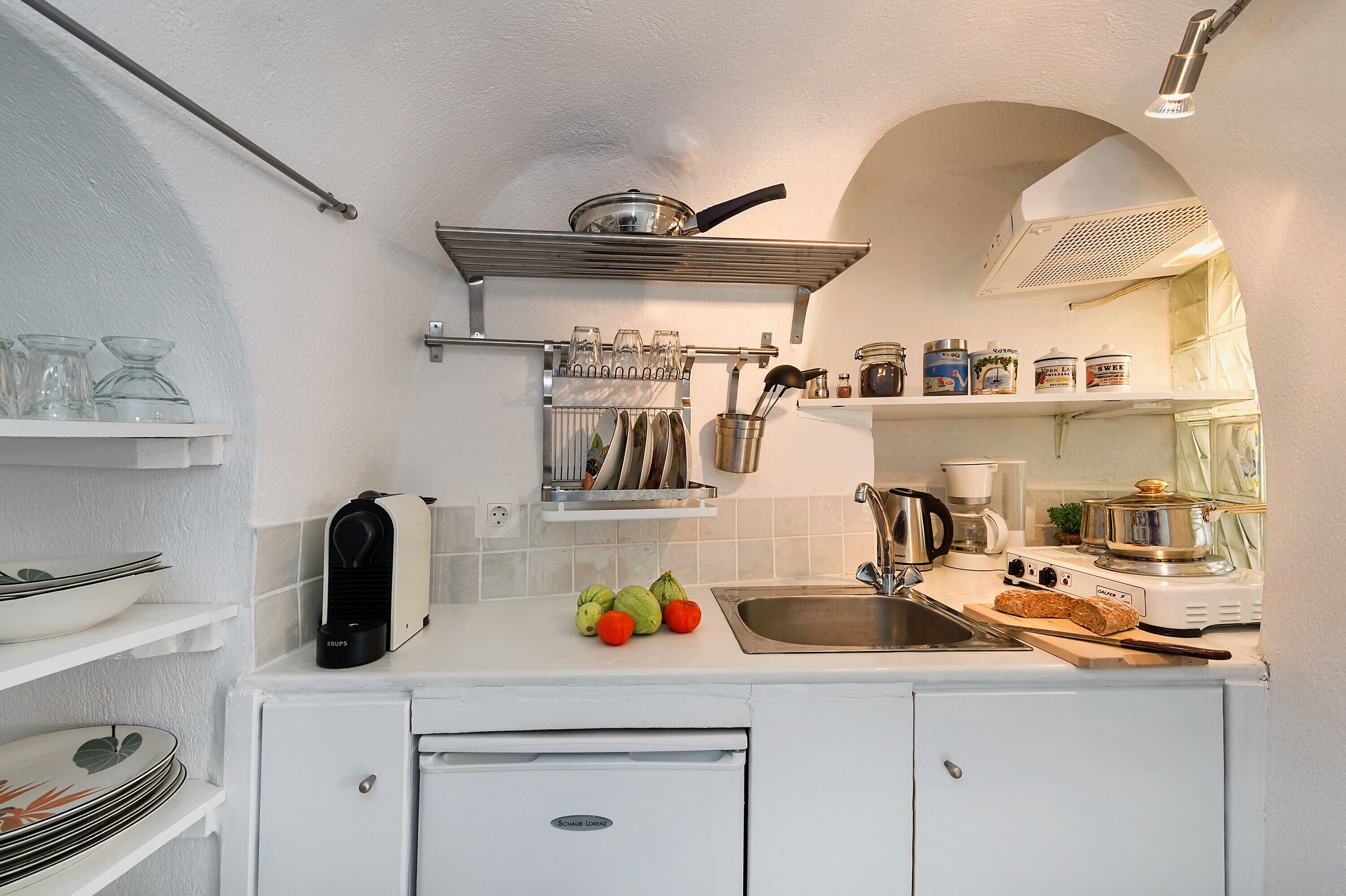 A compact kitchen with white cabinets, a built-in stove, and a sink. There are shelves with glassware and a coffee maker.