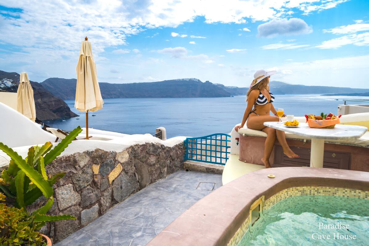 A woman in a bikini sitting at a table with a drink, overlooking a pool and a scenic view of the sea and mountains.