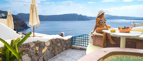 A woman in a bikini sitting at a table with a drink, overlooking a pool and a scenic view of the sea and mountains.