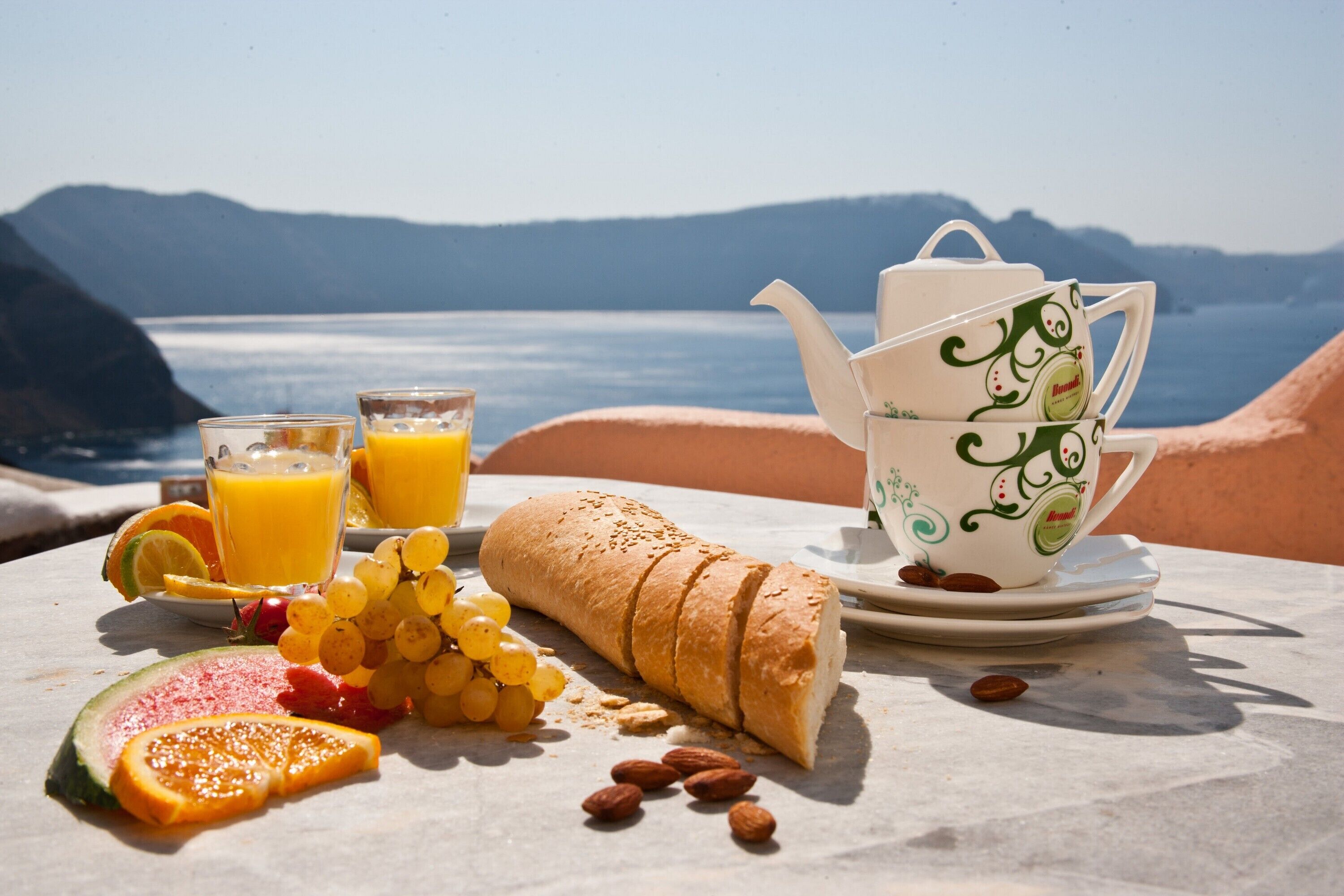 Ontbijt met een croissant, druiven, plakjes watermeloen en sinaasappelsap op een tafel, met op de achtergrond een prachtig uitzicht op bergen en water.