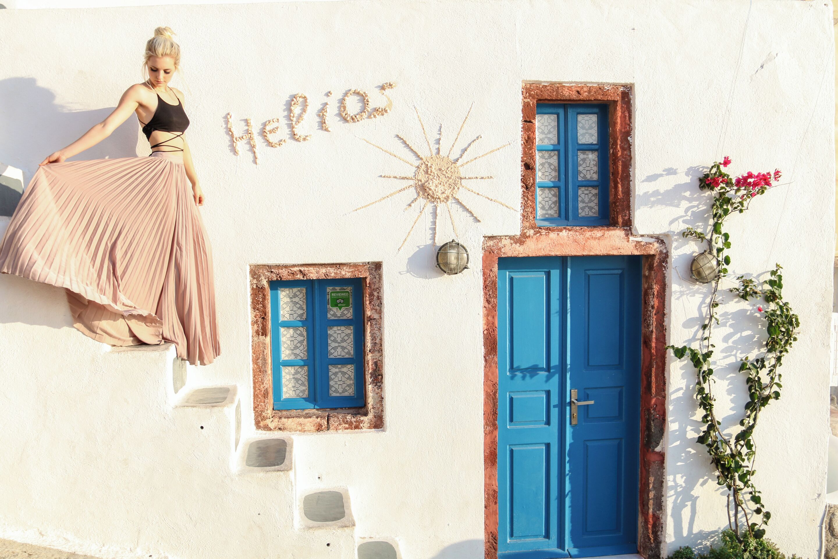 A woman in a pink dress standing on a white building with a blue door and windows, and the word "Helios" written on the wall.