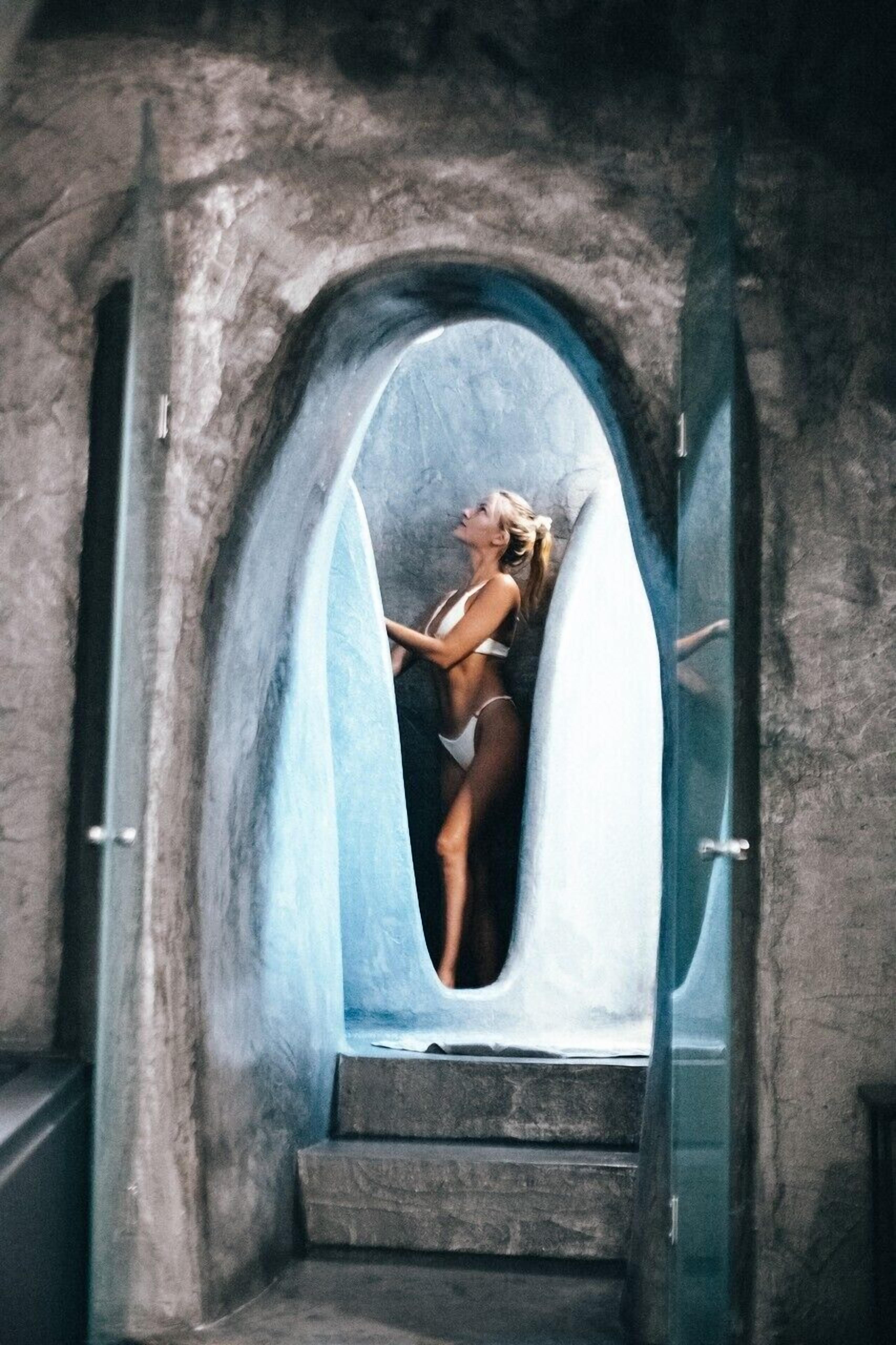 A woman in a bikini standing in a narrow, arched doorway with a surfboard.