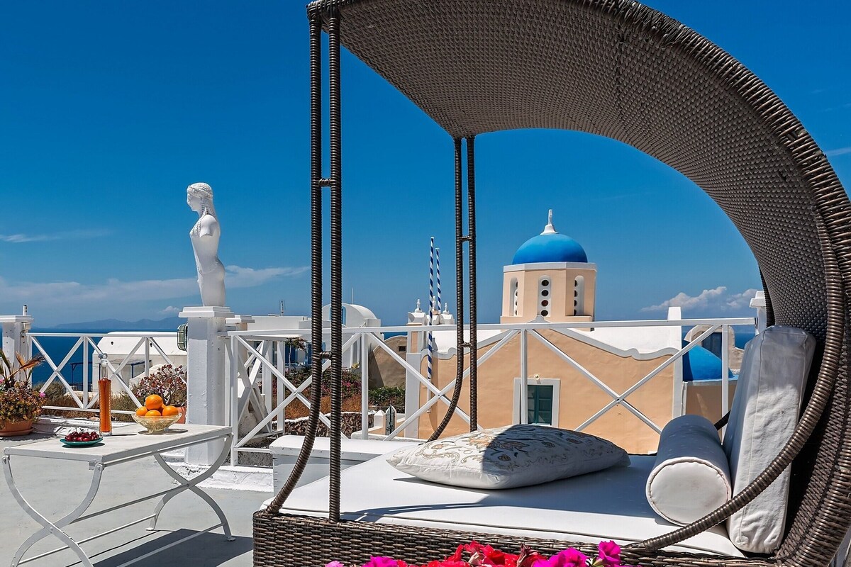 Una silla de mimbre con dosel, una estatua blanca y vistas de un edificio con cúpulas azules.