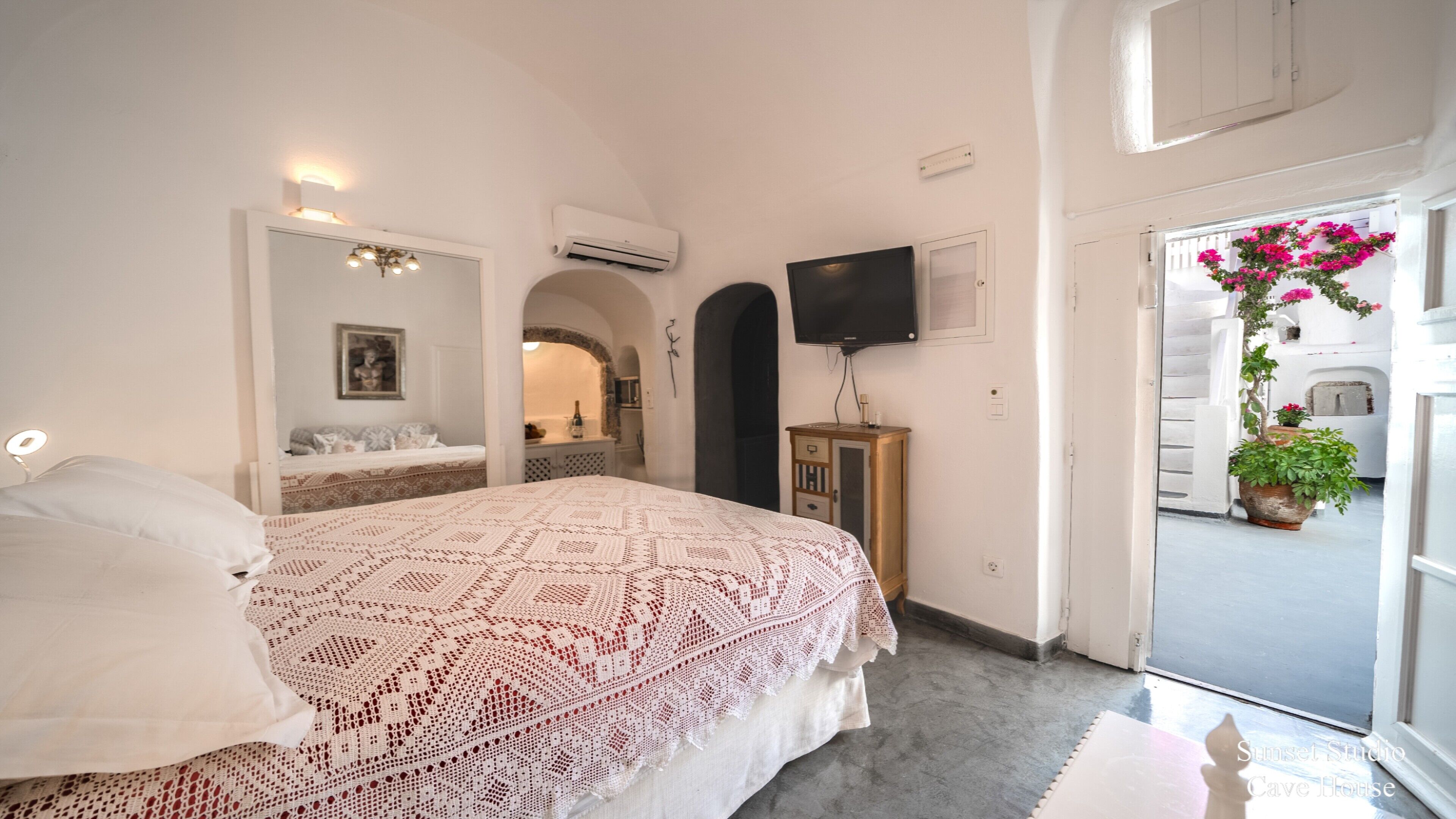 A bedroom with a large bed, a TV, an air conditioning unit, and a view of a white building through an open door.