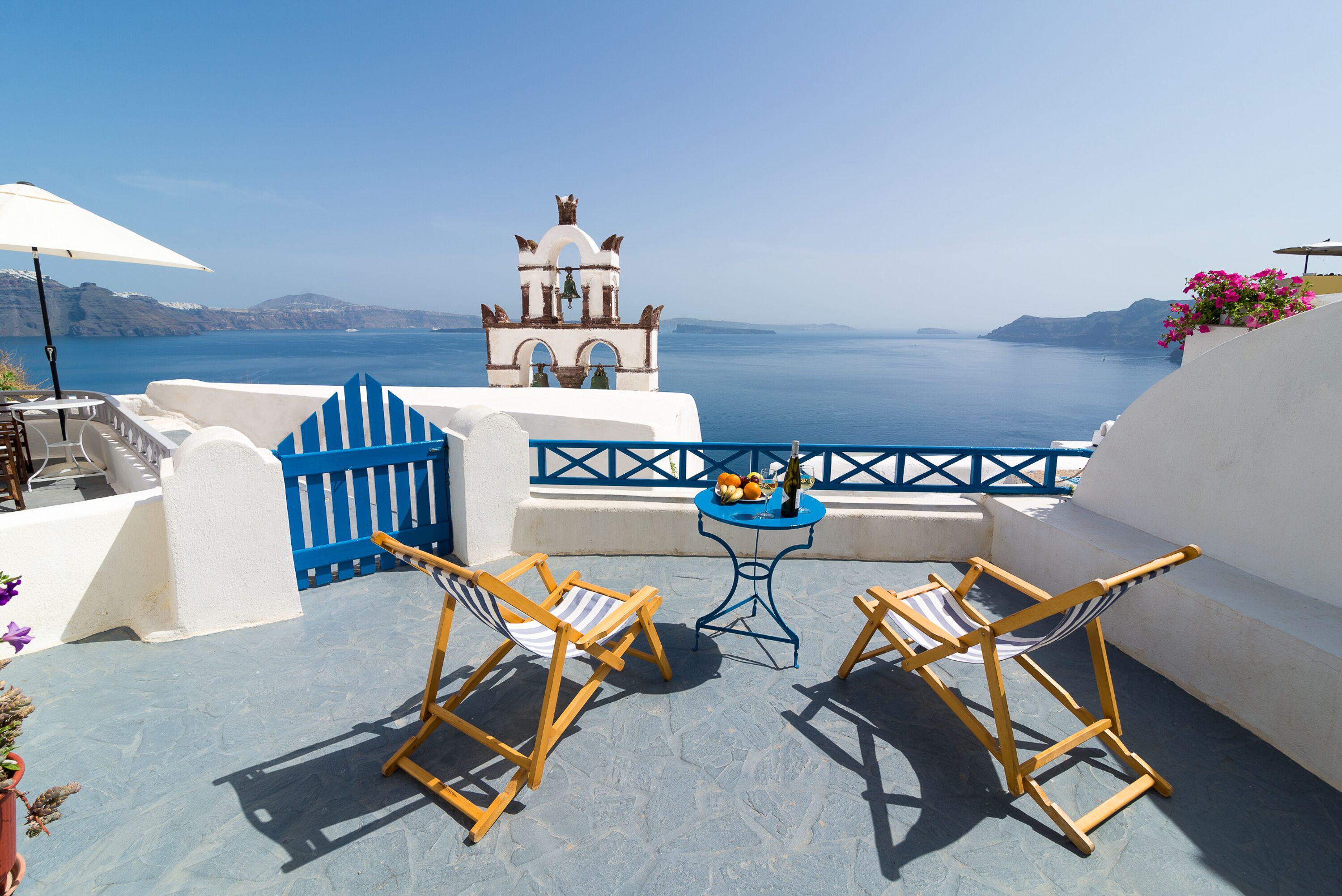 Een terras met twee houten stoelen, een kleine ronde tafel met een fles en fruit, en uitzicht op zee en een klokkentoren.