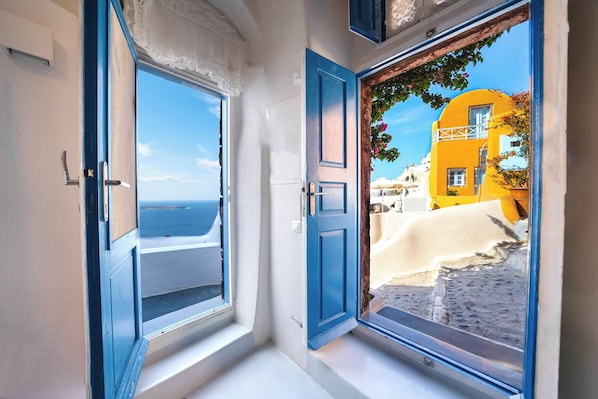 Una habitación con dos ventanas abiertas, una con vista al mar y la otra a un edificio amarillo.