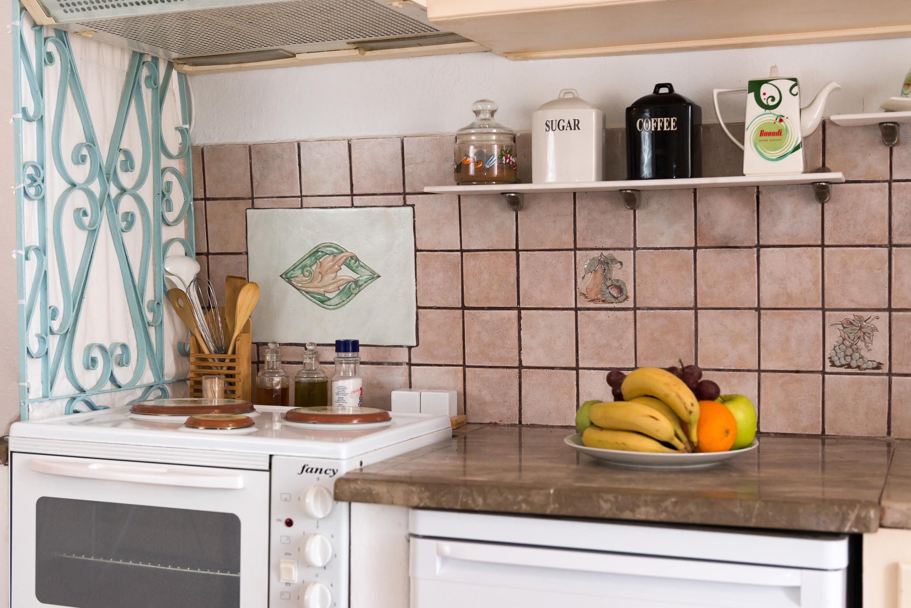 A kitchen with a stove, a sink, and a countertop with a fruit bowl. There are shelves above the countertop holding a sugar container, a coffee container, and a teapot.