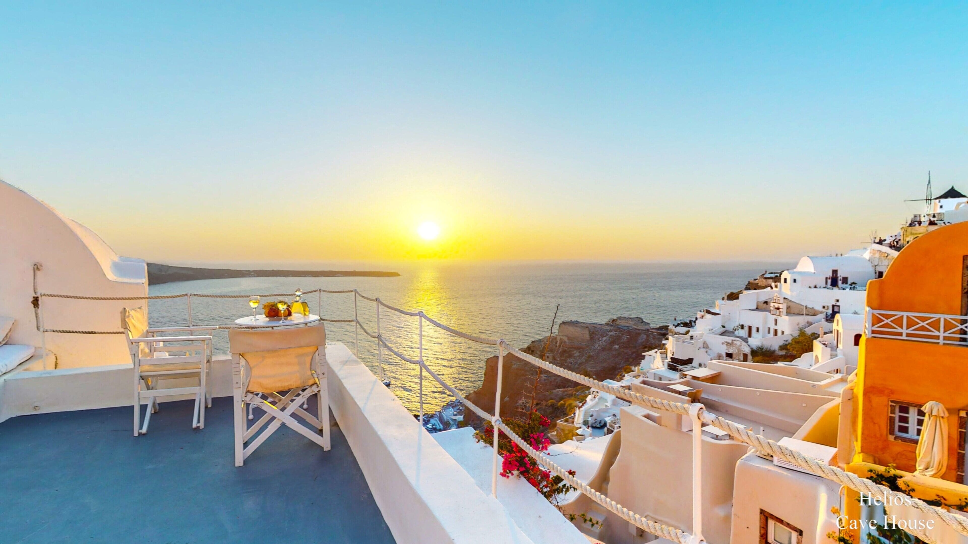 Een balkon met een tafel voor twee, met uitzicht op een kustplaatsje met witte gebouwen en een zonsondergang boven de zee.