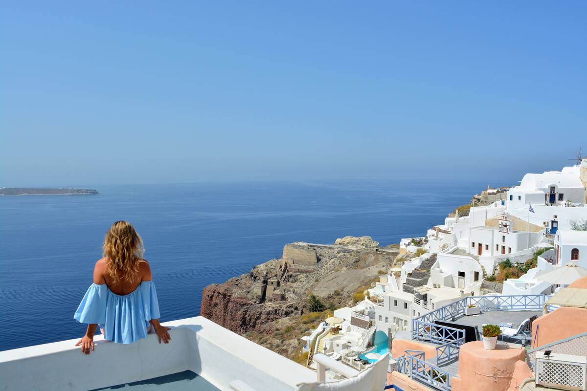 A woman in a blue dress overlooking a coastal town with white buildings and a view of the sea.