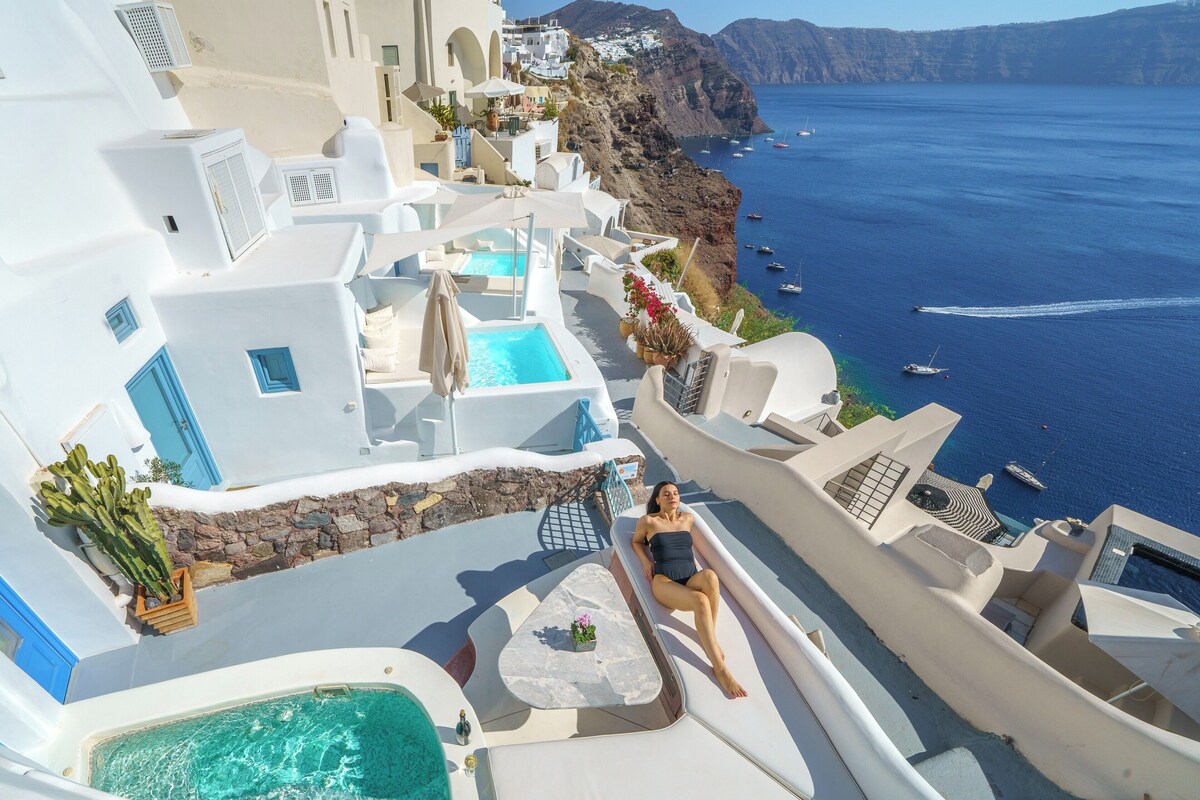 Un edificio blanco con piscina, una mujer relajándose junto a ella, y barcos al fondo.