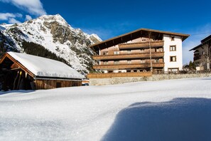 A multi-story wooden building with balconies, surrounded by snow-covered mountains. - Pension Mittagskogel (Sankt Leonhard im Pitztal)