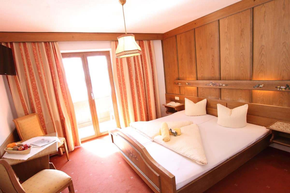 A hotel room with a large bed, wooden headboard, a desk with a chair, a television, and a window with curtains.