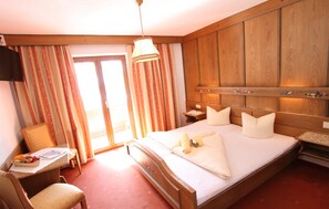 A hotel room with a large bed, wooden headboard, a desk with a chair, a television, and a window with curtains. - Pension Mittagskogel (Sankt Leonhard im Pitztal)