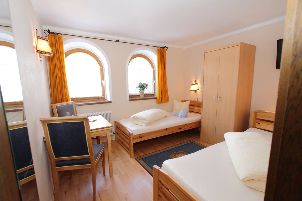 A small, cozy room with a single bed, a desk, and a chair. There is a window with curtains, a radiator, and a wardrobe.
