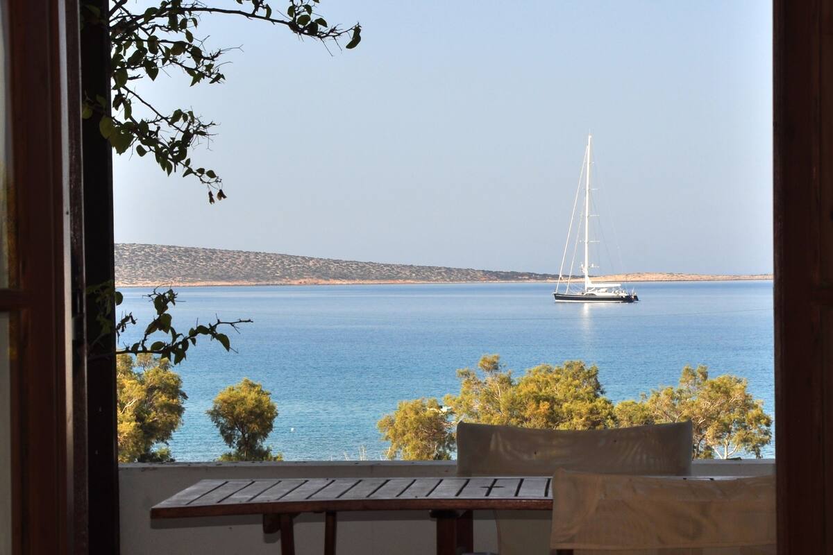Une vue sur une mer calme, avec un voilier, des arbres et une colline au loin.